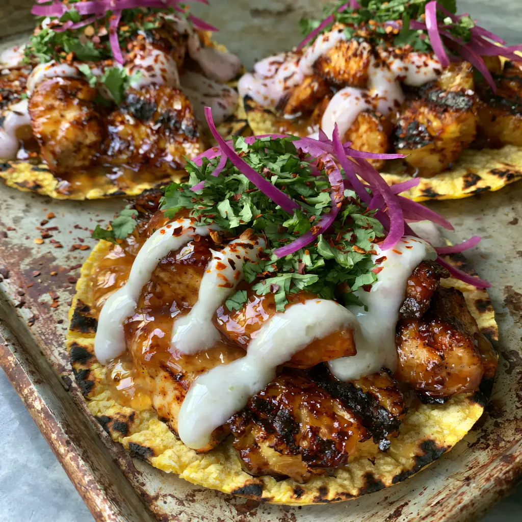 Three pineapple chicken tacos on a seasoned sheet pan showing charred corn tortilla edges and thick lime crema.